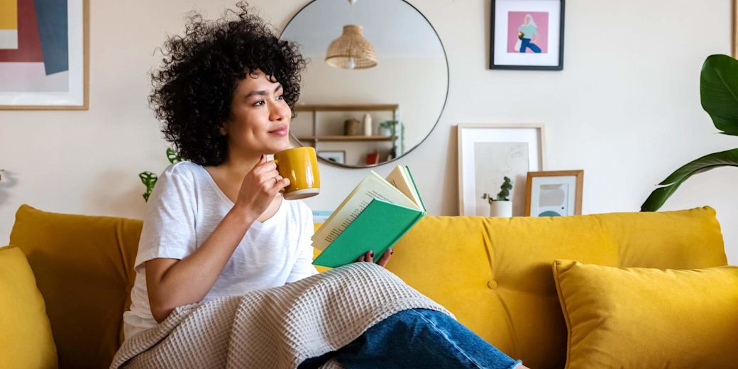 Junge Frau sitzt auf dem Sofa und liest ein Buch mit einer Tasse in der Hand.