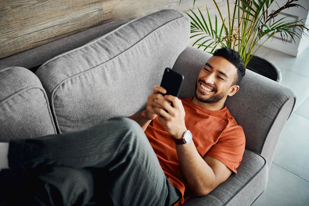 Junger Mann mit orangenem Shirt liegt mit überkreuzten Beinen auf dem Sofa und schaut mit Freude aufs Handy
