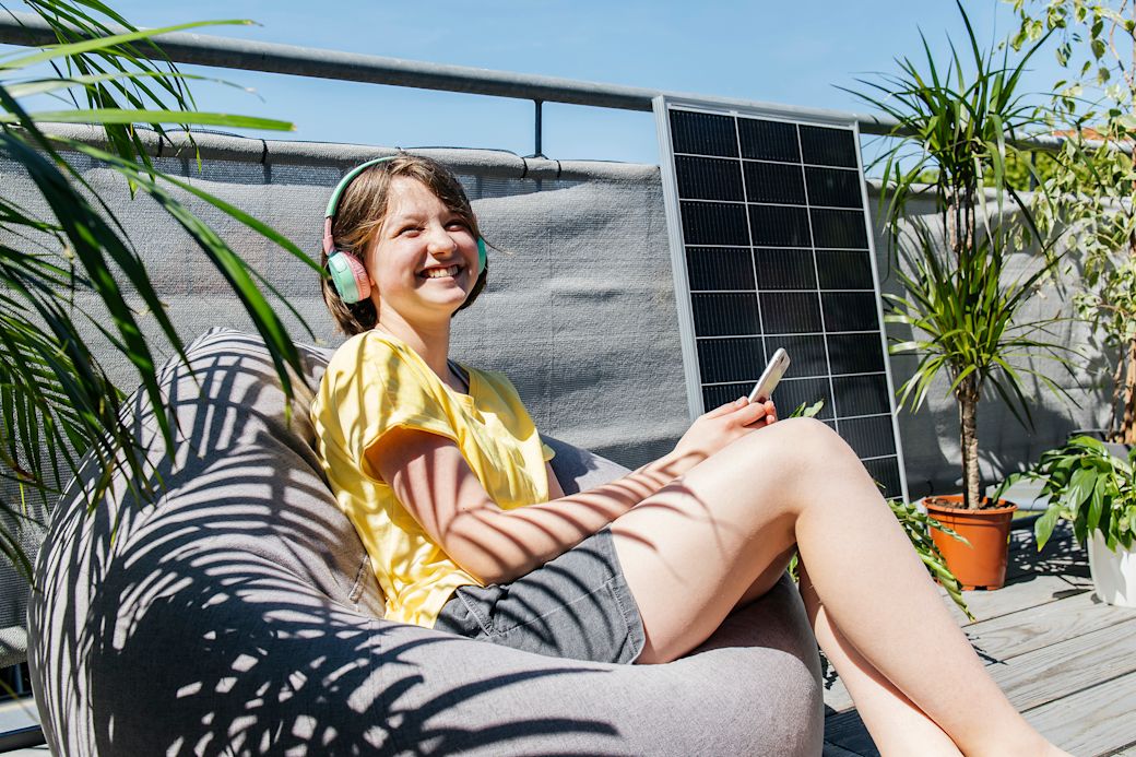 Eine junge Frau sitzt auf ihrem Balkon in der Sonne und hört Musik über ihre Kopfhörer