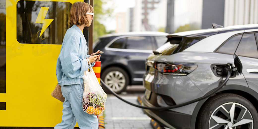 Frau mit Handy und Lebensmitteln wartet auf Laden des Elektroautos.
