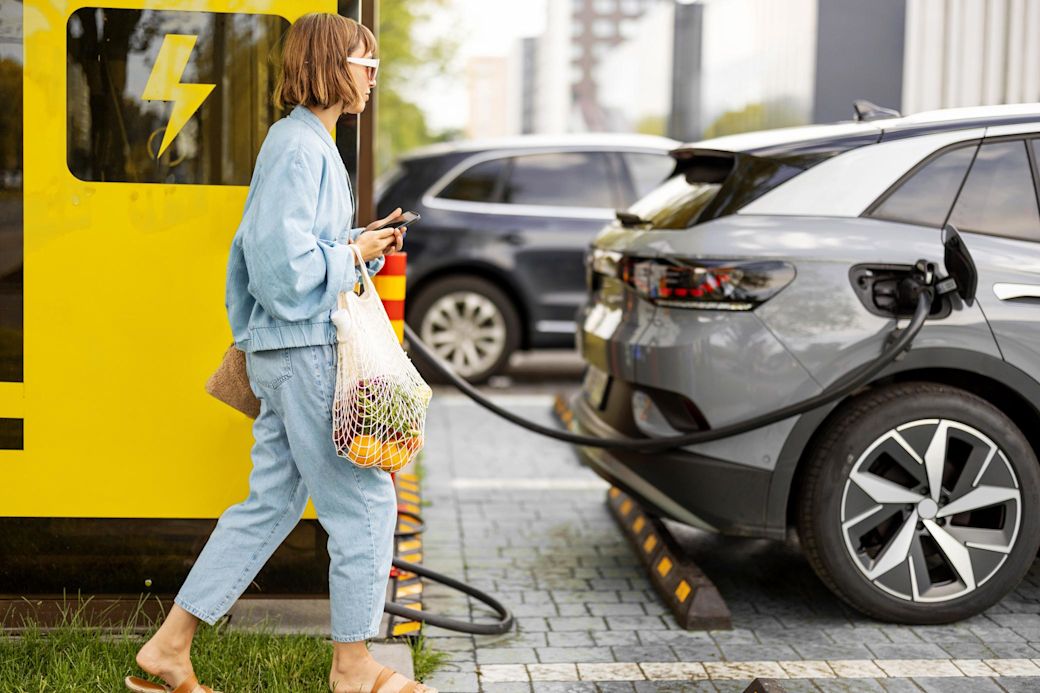 Frau mit Handy und Lebensmitteln wartet auf Laden des Elektroautos