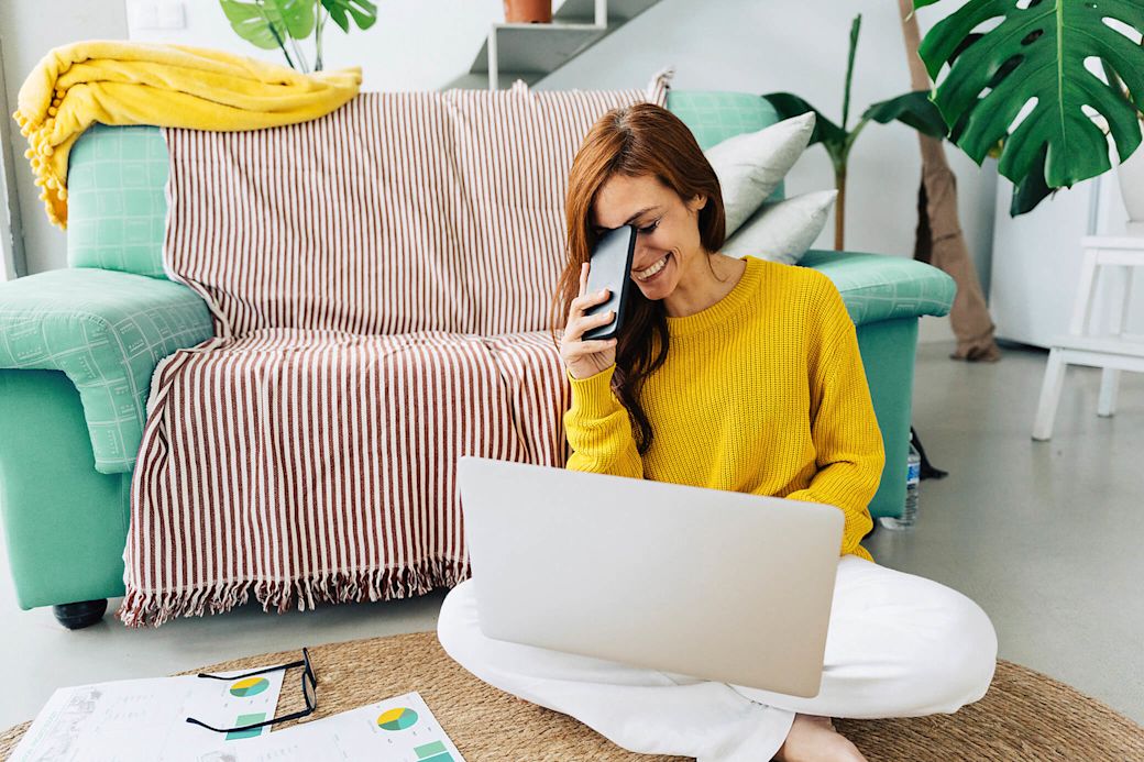 Frau sitzt auf dem Boden mit Handy in der Hand und Laptop auf dem Schoß