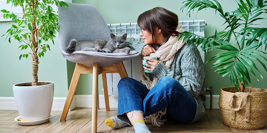 Frau sitzt in warmer Kleidung mit Katze vor der Heizung.