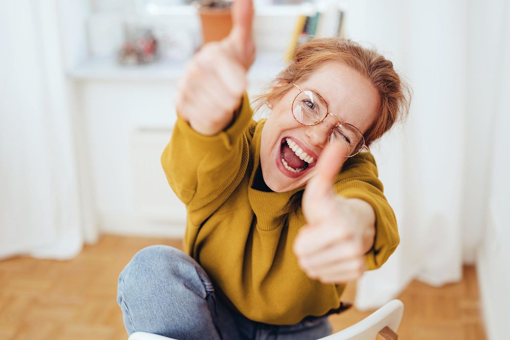 Frau mit Brille und gelbem Pulli zeigt happy beide Daumen hoch in ihrer Wohnung 