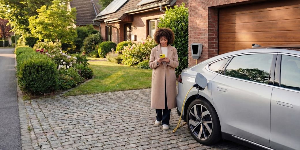 Fröhliche Frau lädt ihr graues E-Auto Zuhause über die Wallbox und steuert Smart Charging intelligent per Handy in der Hand in der Einfahrt vor ihrem grünen Garten
