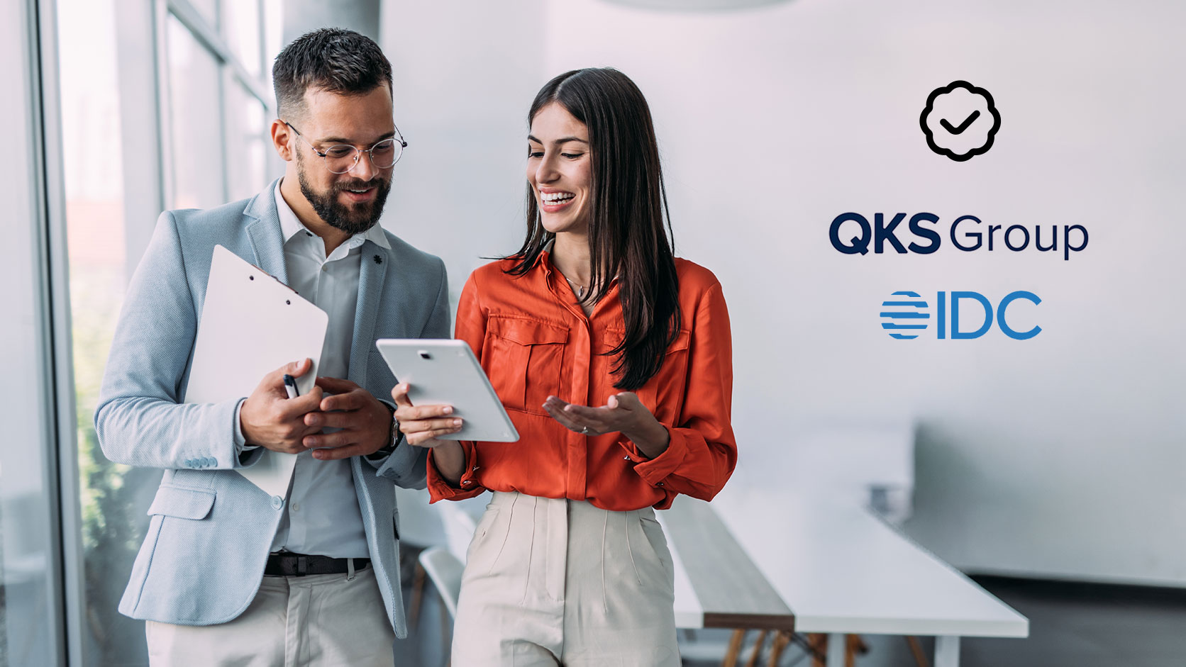 Two business professionals in an office reviewing tablets together, with QKS Group and IDC logos displayed on the right.