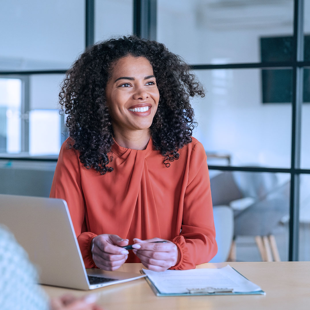 Une femme souriante aux cheveux noirs bouclés, vêtue d’un chemisier orange, est assise à un bureau avec un ordinateur portable dans un cadre de bureau moderne.