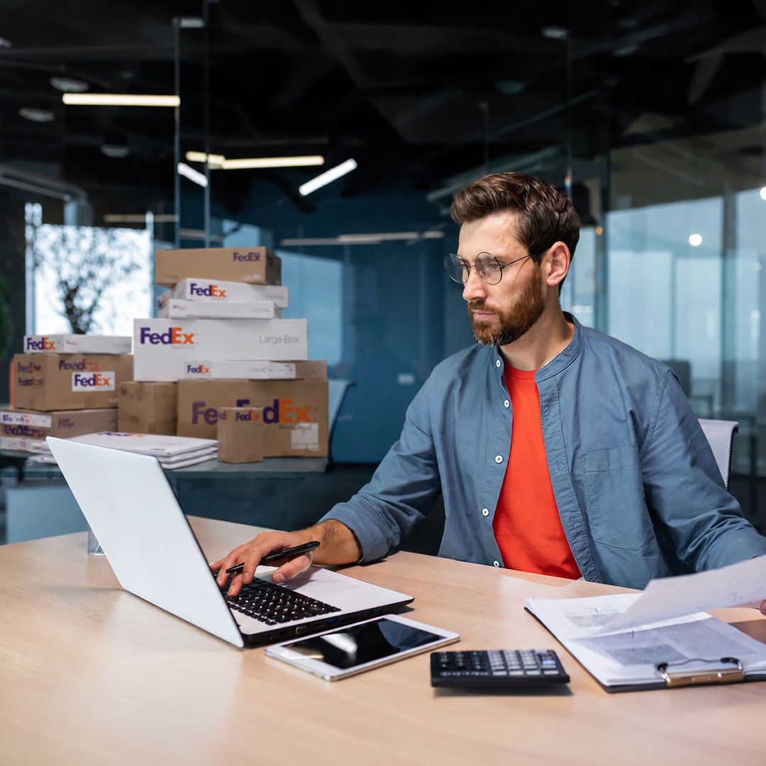 Man in blue shirt and glasses working on laptop at desk with FedEx boxes stacked in modern office background.