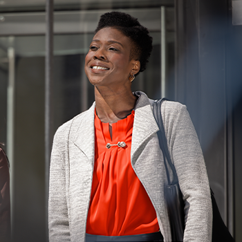 Smiling professional woman in orange blouse and gray blazer with bag strap, standing outdoors near building.