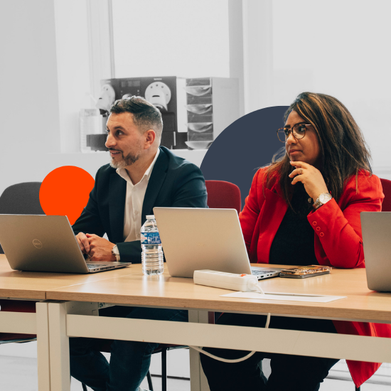 Deux professionnels d’affaires assis à une table de conférence avec des ordinateurs portables, une bouteille d’eau et un cahier dans un cadre de bureau moderne.