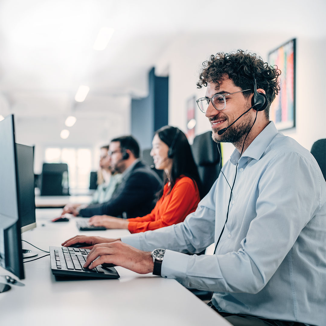 Smiling customer service representative with headset and glasses working at computer in modern office call center.