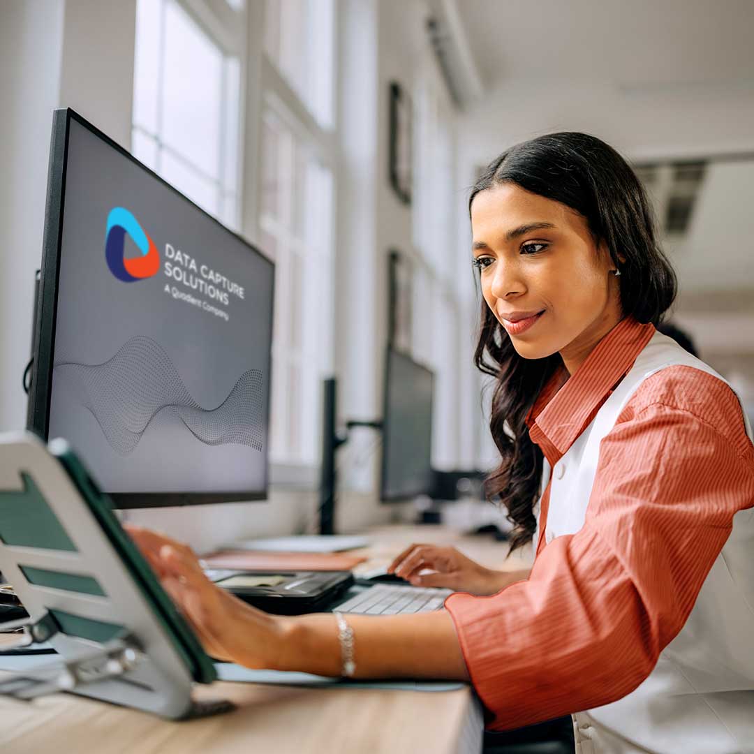Woman in coral and white blouse working at computer displaying Data Capture Solutions logo in bright office setting.