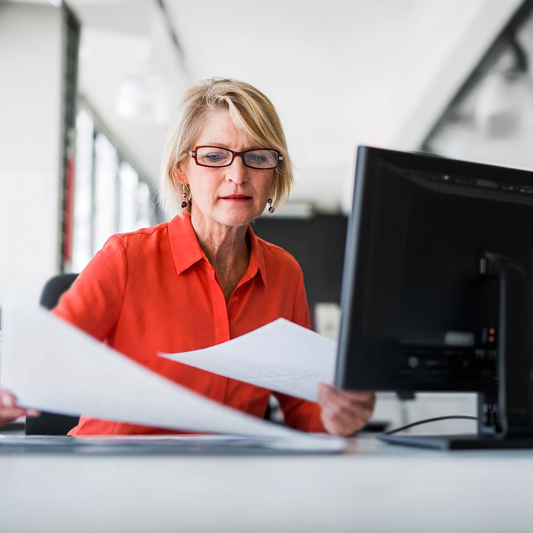 Woman with blonde hair and glasses wearing an orange shirt reviews documents at a desk with a computer monitor.