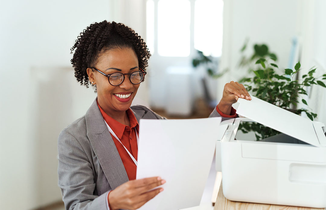 Smiling businesswoman in gray blazer and red blouse using office printer to retrieve printed documents in bright office.
