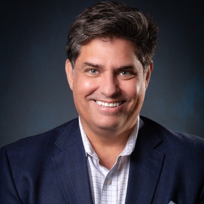 Professional headshot of a dark-haired man wearing a navy blazer and light shirt, facing forward against a plain dark background.