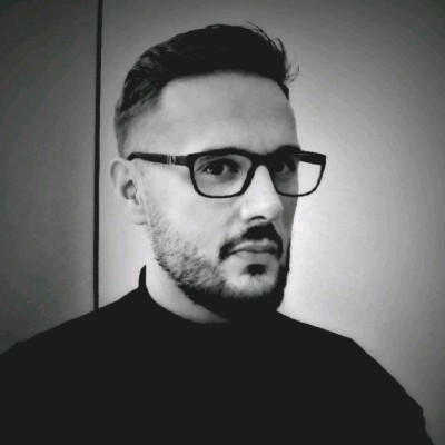 Black-and-white portrait of a man with short hair and a trimmed beard, wearing rectangular glasses and a dark high-collar top, facing slightly to the side against a plain, softly lit background.