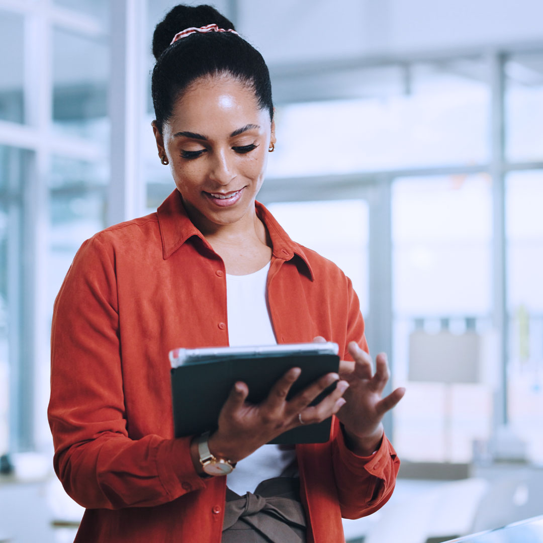 Woman in orange shirt with hair in bun smiling while using tablet in bright modern office setting
