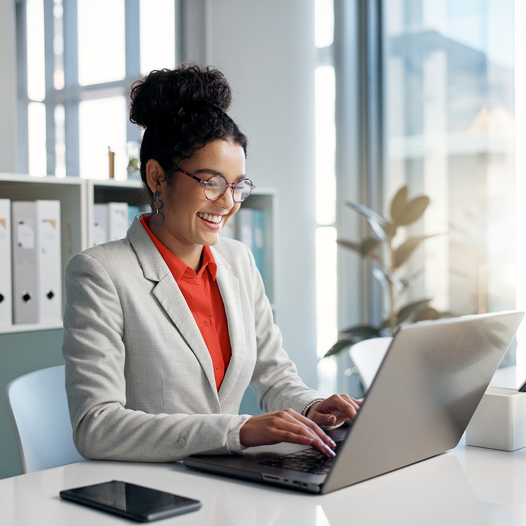 Smiling businesswoman in gray blazer and red shirt working on laptop in bright modern office with windows.