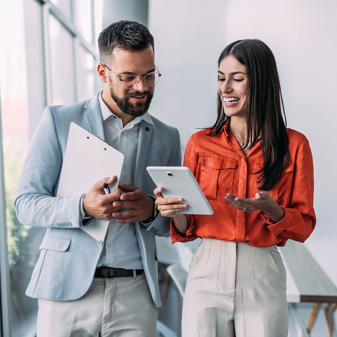 Two business professionals in a modern office reviewing content on a tablet together, man in gray suit and woman in orange blouse.
