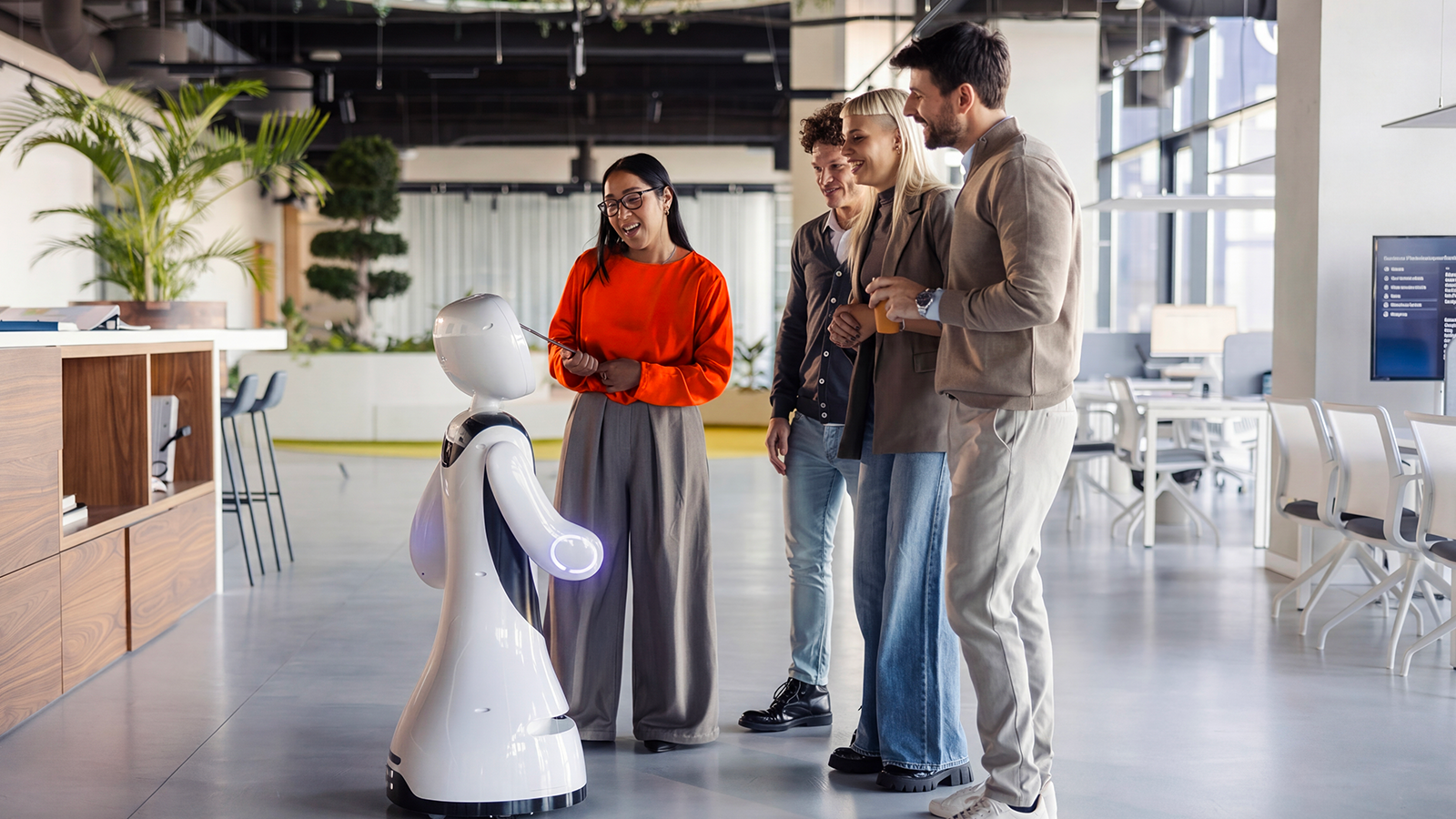 Woman in orange sweater demonstrating white humanoid robot to three colleagues in modern office with plants and windows.