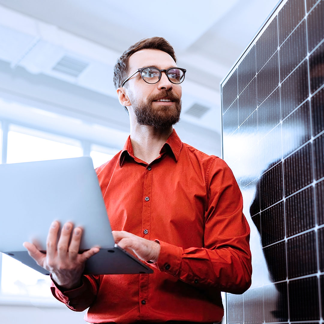 Man in red shirt and glasses, potential utilities expert, holding laptop standing next to solar panel in bright modern office setting.