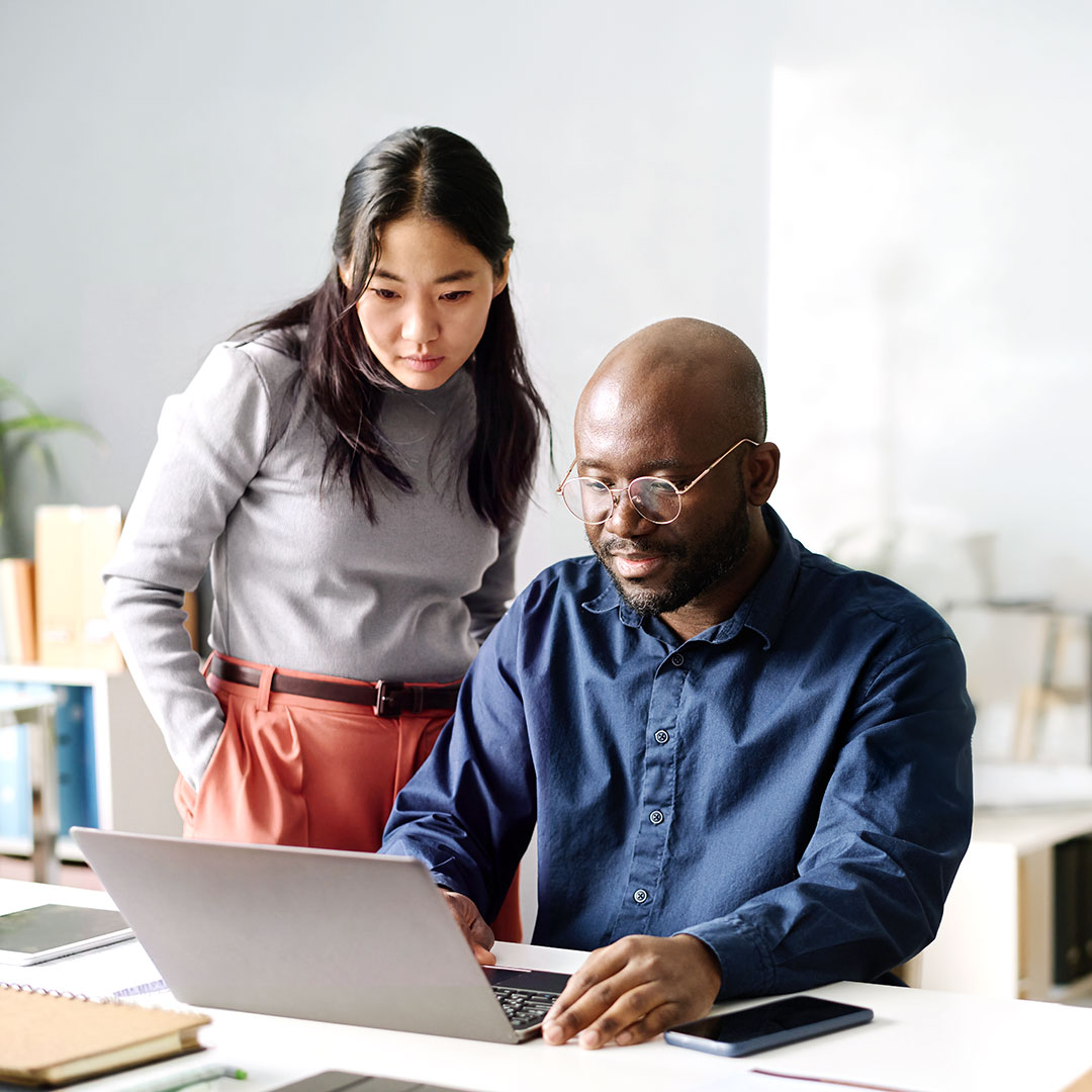Asian woman in gray sweater and coral pants standing beside Black man in blue shirt working on laptop in office.