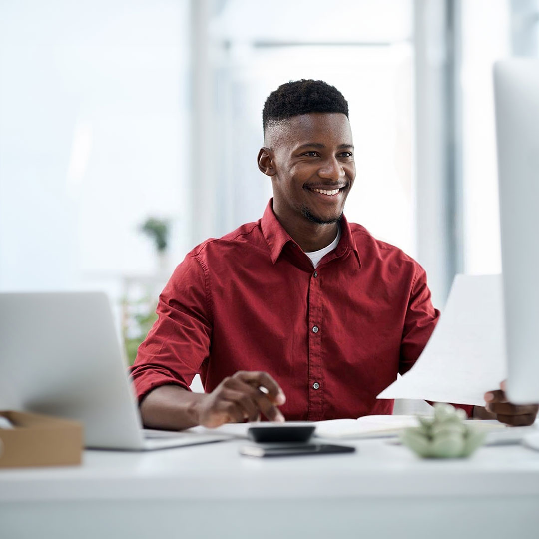 Smiling man in red shirt working at desk with laptop and documents in bright modern office setting