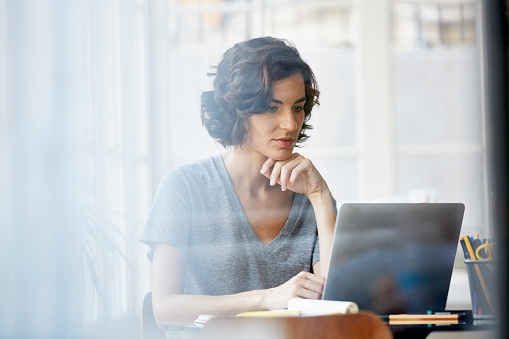 A woman sits at a table working on a laptop, looking focused as she rests her chin on her hand. Soft daylight filters through the window in front of her.