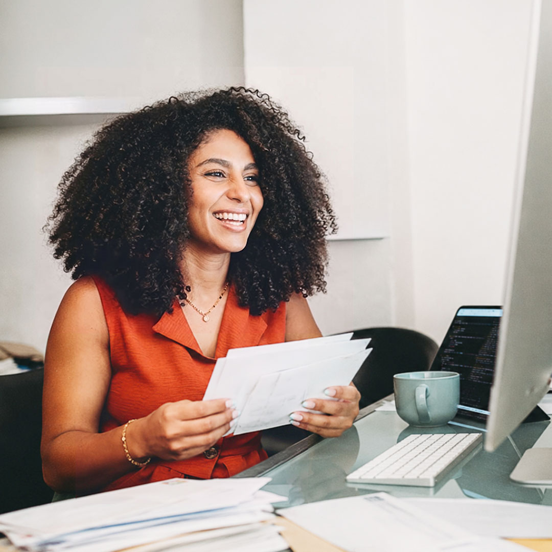 Smiling woman with curly hair in orange dress holding envelopes and mail at desk with computer and coffee cup in bright office.