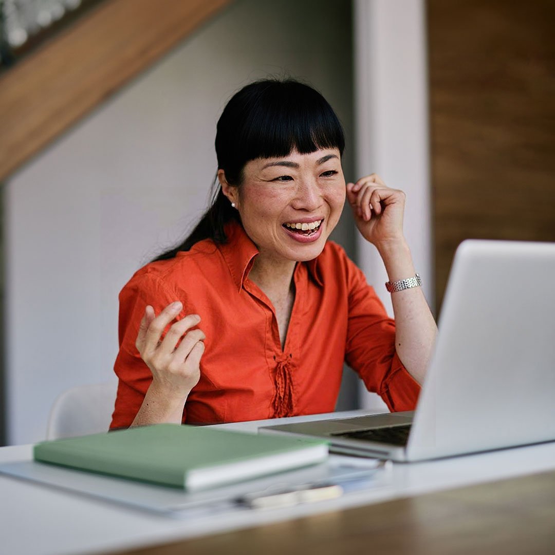 Woman with black hair wearing orange shirt laughing while gesturing during video call at desk with laptop and notebook.
