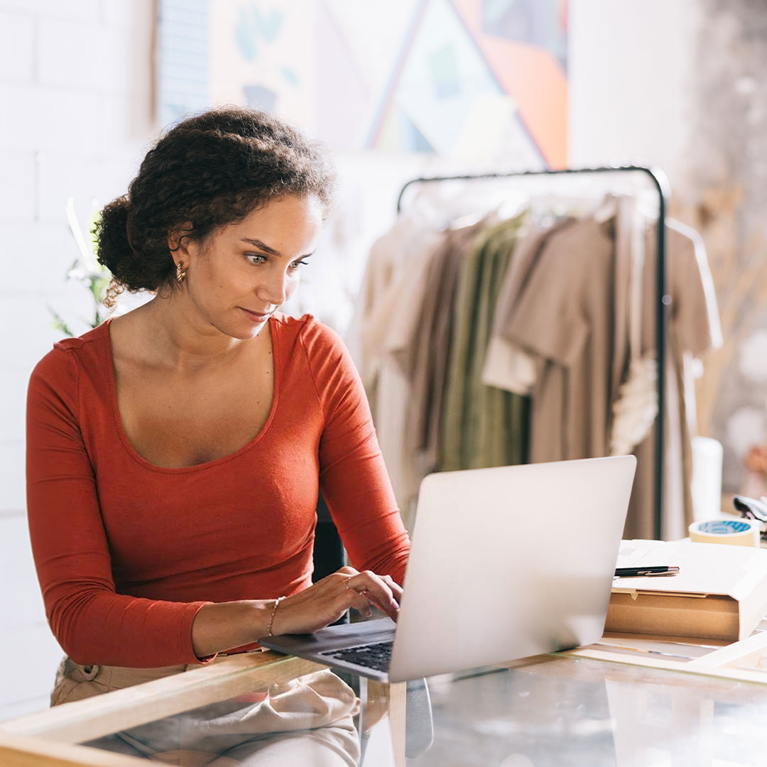 Woman in orange top working on laptop at desk with clothing rack in background in bright retail or studio space.