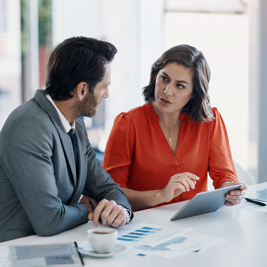 Woman in red blouse and man in gray suit discussing business with tablet and documents at office table.