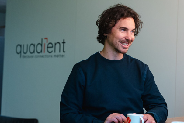 un jeune homme tenant une tasse avec le logo Quadient et portant un chandail bleu foncé, souriant en regardant à droite