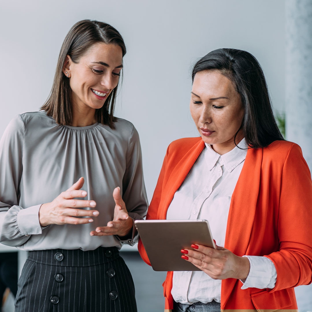 Two professional women collaborating, one pregnant in gray blouse, one in orange blazer holding tablet, smiling together.