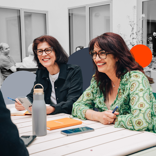 Two smiling women with glasses sit at a white table with notebooks and water bottles during a meeting in a bright office.