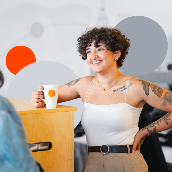 Smiling woman with curly hair and tattoos holding a coffee mug at a wooden table in a modern office setting.