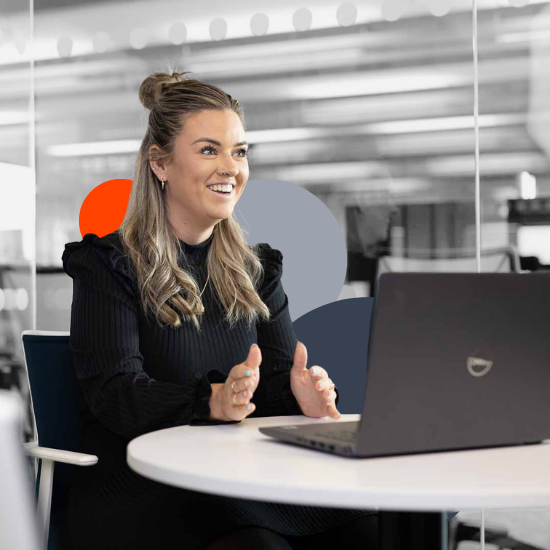 Smiling woman with blonde hair in black sweater gesturing while sitting at laptop in modern office