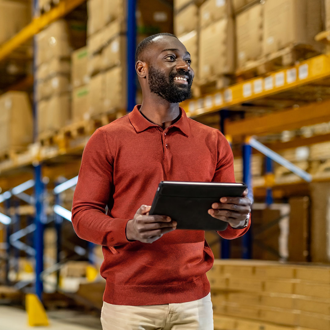 Smiling warehouse worker in red polo shirt holding tablet, standing among shelves stocked with cardboard boxes.