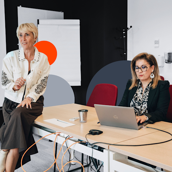 Two professional women in a modern office meeting room, one standing presenting, one seated at laptop with colorful wall art