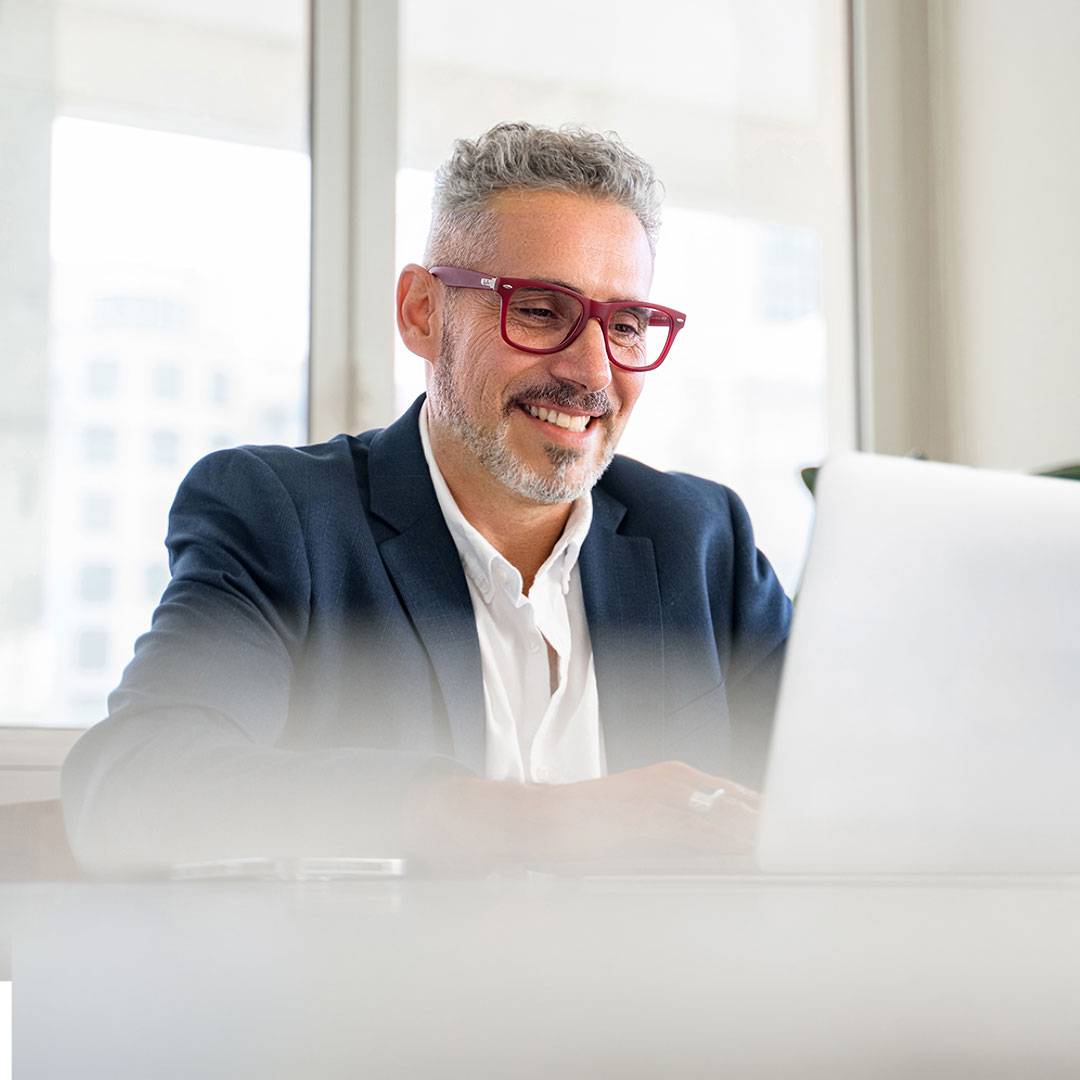 Smiling businessman with gray hair and red glasses wearing a navy suit working on laptop in bright office.