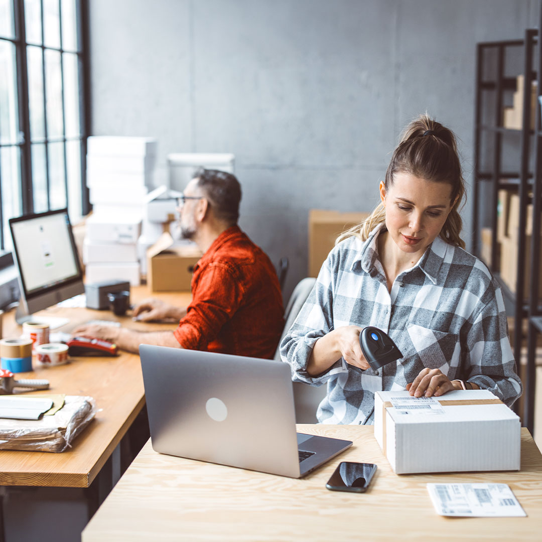 Woman in plaid shirt scanning package label at desk with laptop while man works at computer in background office.