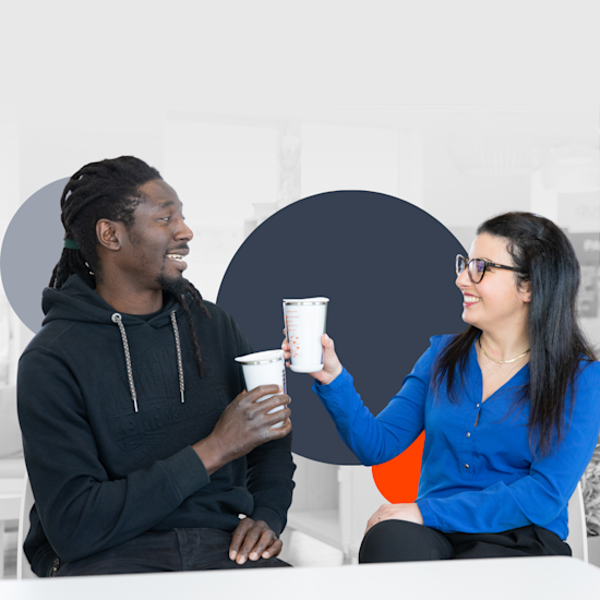 Two coworkers having coffee together in a modern office, man in black hoodie and woman in blue shirt smiling and chatting