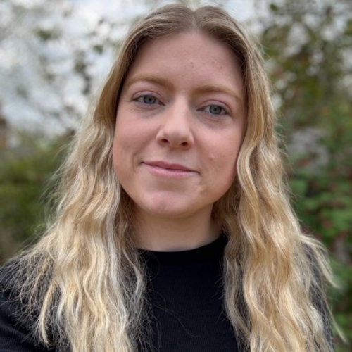 Outdoor headshot of a blond woman with long wavy hair, wearing a black top and a slight smile, against a blurred natural background.