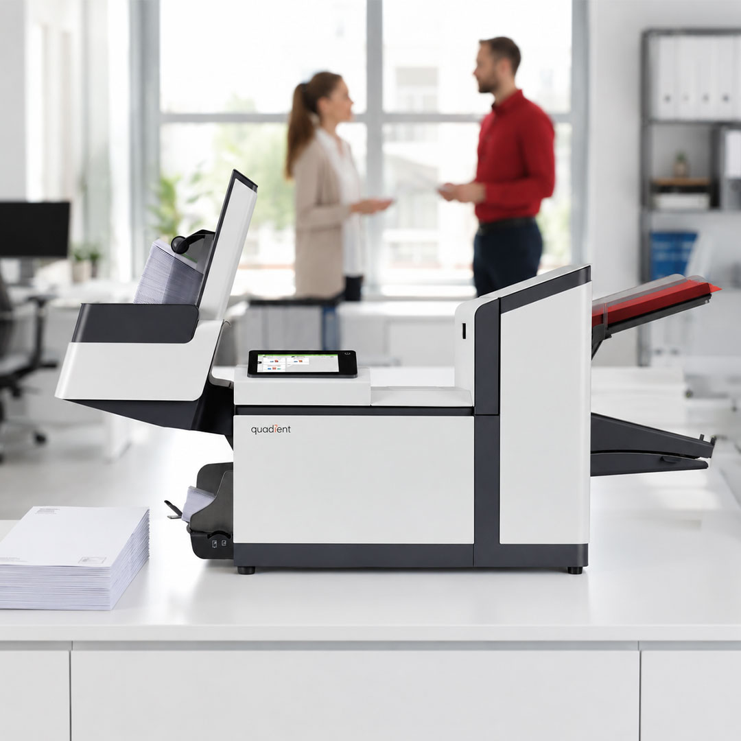 Quadient mail folder inserter machine on white desk with two professionals conversing in bright modern office background.