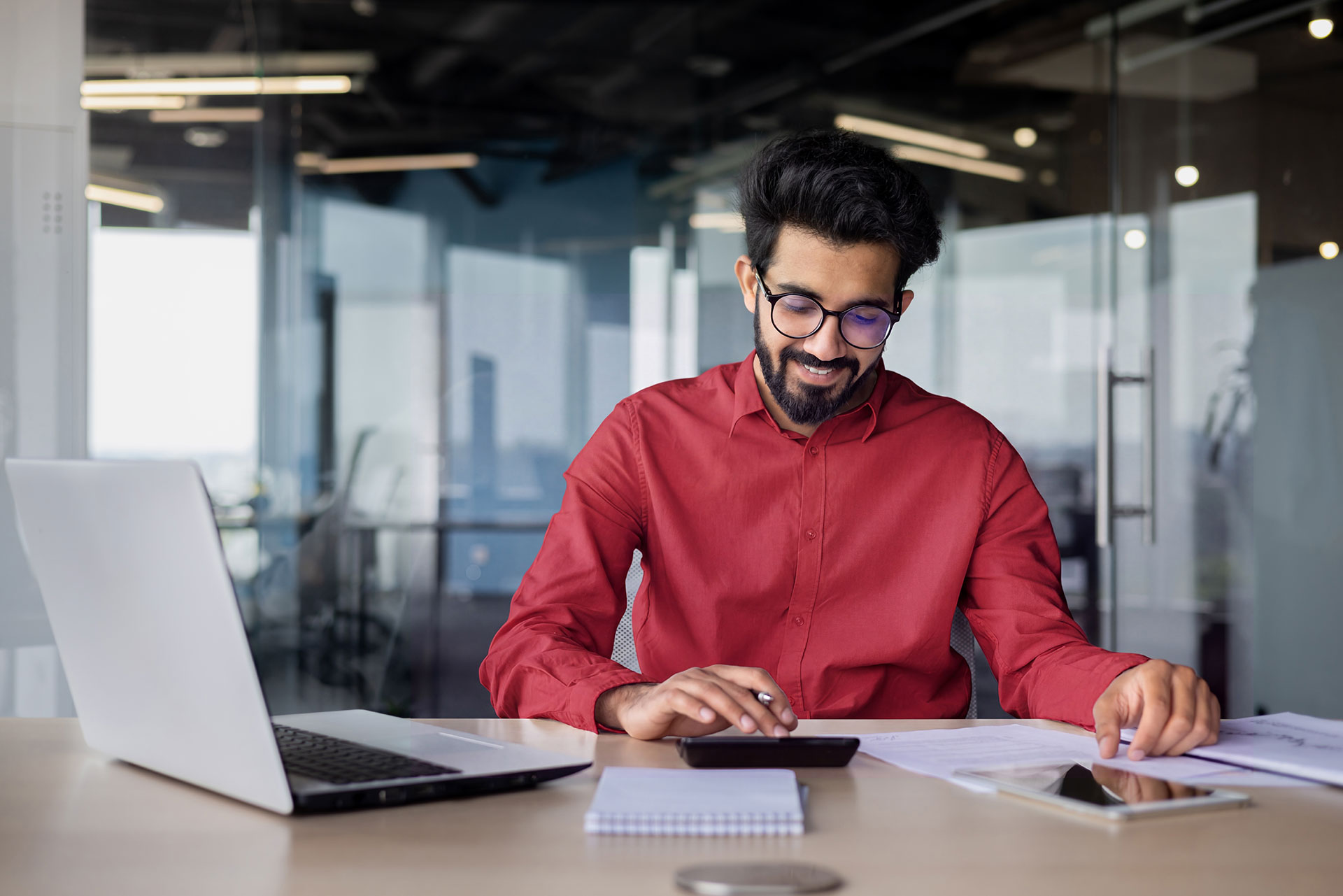 Man in red shirt and glasses working at desk with laptop, calculator, and documents in modern office setting.