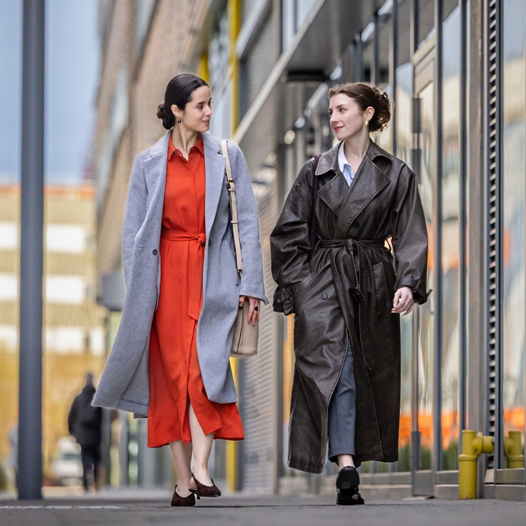 Two women walking together on an urban street, one wearing a gray coat with orange dress, the other in a black leather coat.