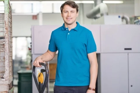 Smiling man in blue polo shirt holding vacuum cleaner in bright modern office space with white walls and shelving.