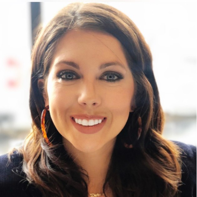 Close-up portrait of a woman with shoulder-length brown hair, wearing large hoop earrings, facing the camera with a smile. Soft indoor lighting and a blurred background.