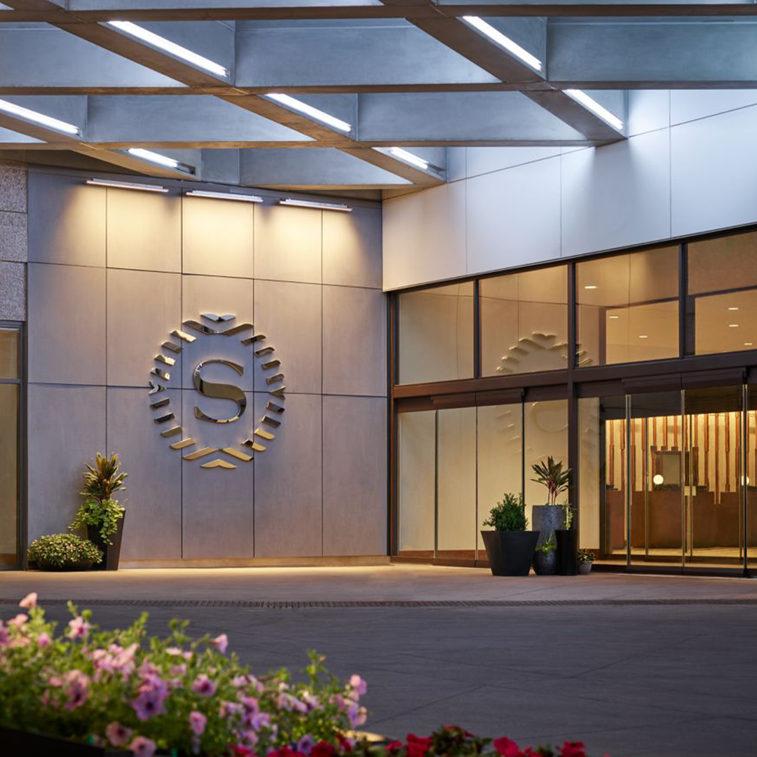 Modern hotel entrance with illuminated 'S' logo on beige wall, glass doors, wooden beams, and decorative plants at dusk.