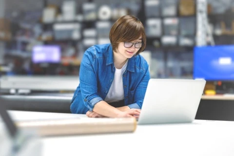 Woman in blue shirt and glasses working on laptop at desk in modern office with monitors in background.